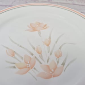 May include: White ceramic plate with a floral design. The plate features peach-colored flowers and buds with green stems and leaves. A peach and green border encircles the rim.