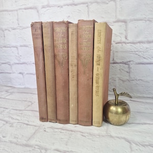 May include: A collection of antique books with titles including "Librettos of the French and German Operas" and "Brettos of the Italian Operas." The books are in various shades of pink and beige. A small brass apple decorates the scene.
