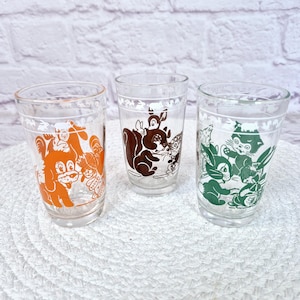 May include: Three vintage clear drinking glasses with cartoon animal designs in orange, brown, and green. The glasses are cylindrical and sit on a white woven mat. The designs include a dog, squirrel, and cat.