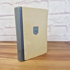 May include: A vintage book with a light tan cover and a blue spine. The spine has text and the cover features a blue emblem. The book is standing upright on a wooden surface.