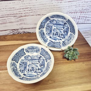 May include: Two small, white ceramic bowls with a blue and white transferware design depicting a detailed street scene. The bowls have a scalloped edge and are displayed on a wooden surface.