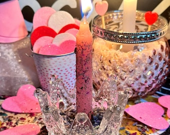 Puede incluir: Una vela rosa con un portavelas de vidrio en forma de corona se encuentra sobre una mesa con corazones de fieltro rosa, lavanda seca y pétalos de rosa. La vela está encendida y la llama es visible.