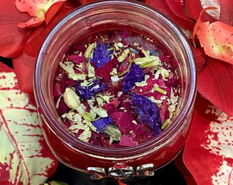 Puede incluir: Una vela de vidrio rojo en un frasco con un diseño floral rojo, blanco y morado. La vela está rodeada de pétalos de flores rojas.