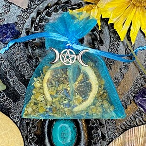 May include: A blue tulle pouch with a silver pentagram charm and dried herbs and a lemon slice inside. The pouch is on a black background with other crystals and flowers.