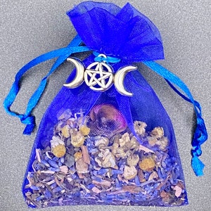 Op de afbeelding: Een blauwe gaas zak gevuld met gedroogde kruiden en bloemen, vastgebonden met een blauw lint en versierd met een zilveren drievoudige maan en pentagram bedel.