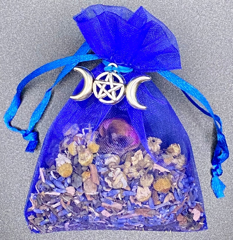 Op de afbeelding: Een blauwe gaas zak gevuld met gedroogde kruiden en bloemen, vastgebonden met een blauw lint en versierd met een zilveren drievoudige maan en pentagram bedel.