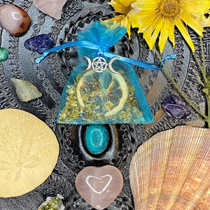 May include: A blue tulle pouch with a dried lemon slice, herbs, and a silver triple moon pendant with a pentacle charm. The pouch is surrounded by various crystals, seashells, and a small bottle labeled "Witch Sage".