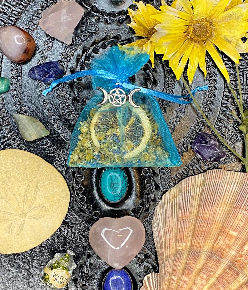 May include: A blue tulle pouch with a dried lemon slice, herbs, and a silver triple moon pendant with a pentacle charm. The pouch is surrounded by various crystals, seashells, and a small bottle labeled "Witch Sage".
