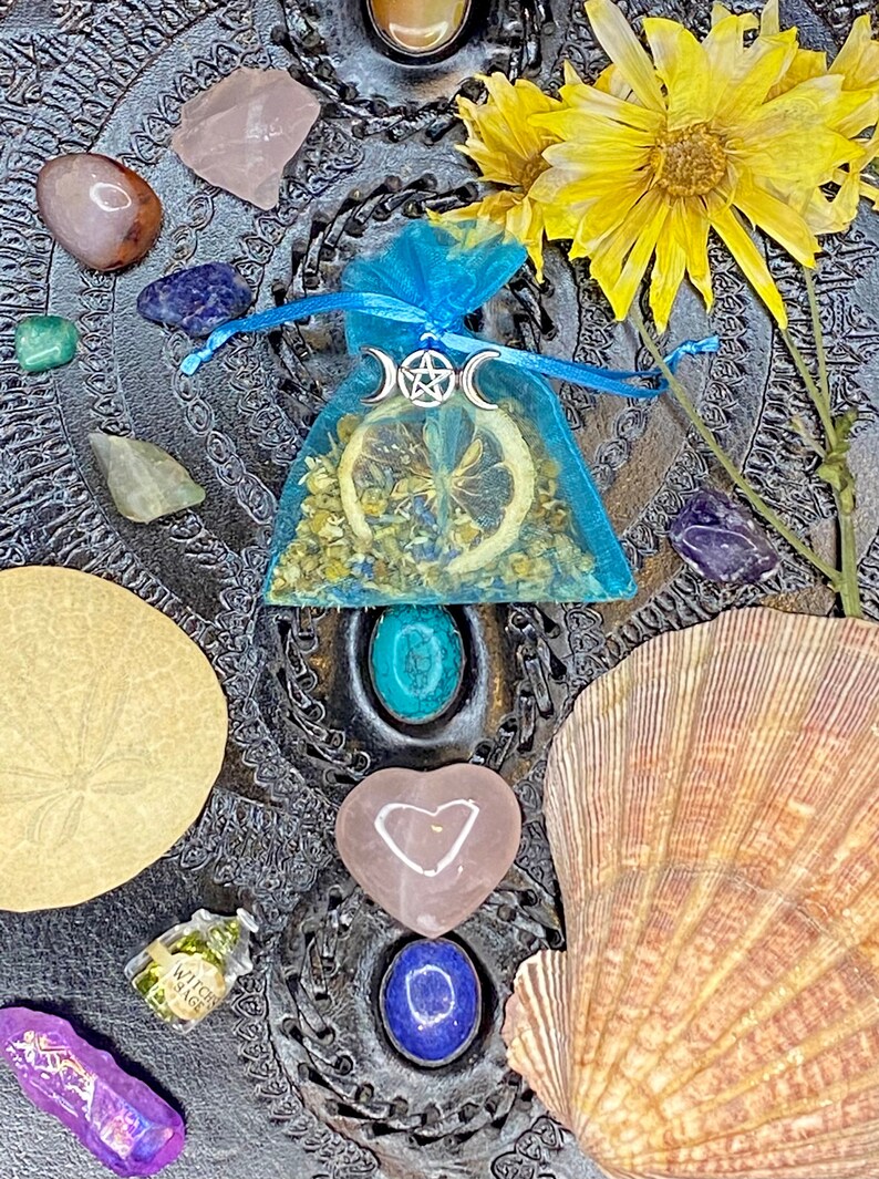 May include: A blue pouch with a dried lemon slice and dried flowers inside. The pouch has a silver charm with a triple moon and pentacle symbol. The pouch is on a black surface with various crystals, a seashell, and a small bottle labeled "Witch Sage".