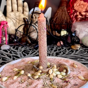 May include: A lit, textured, dusty pink candle stands in a small, circular, ceramic candle holder. The holder sits in a shallow, white, round dish filled with pink salt, scattered dried flower buds, and small herbs, creating a layered display.