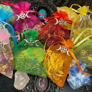 May include: Assortment of colorful, sheer fabric pouches filled with dried herbs and flowers. Each pouch features a silver charm with a triple moon and pentacle design. The pouches are arranged on a dark surface with various crystals.