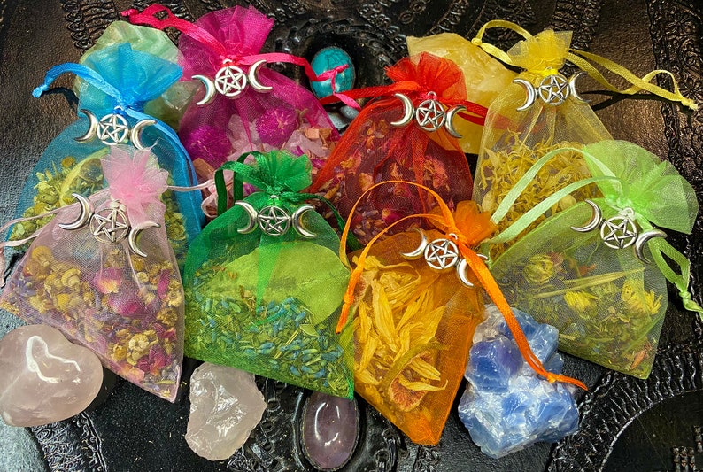 May include: Assortment of colorful, sheer fabric pouches filled with dried herbs and flowers. Each pouch features a silver charm with a triple moon and pentacle design. The pouches are arranged on a dark surface with various crystals.