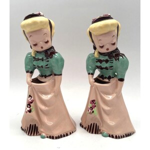 May include: A pair of vintage ceramic planter figurines of girls in dresses with floral accents. The girls are holding their skirts up to reveal a planter inside.