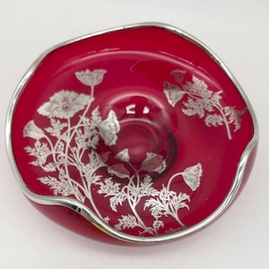 May include: A circular, deep red glass bowl featuring a metallic silver floral pattern of poppies across its interior surface. The bowl has an irregular, wavy rim finished with a smooth silver-colored metallic edge that follows the contour.
