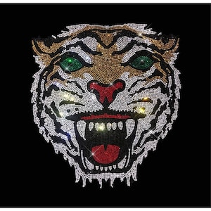May include: A close-up of a tiger's face made of rhinestones. The tiger has a fierce expression with its mouth open and teeth showing. The rhinestones are clear, gold, red, and green.