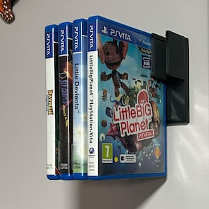 May include: A wall-mounted video game storage rack with four blue video game cartridges. The cartridges are for the PlayStation Vita and include the games LittleBigPlanet, Little Deviants, and Rayman.