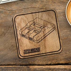 May include: Square wooden coaster with a laser-etched design of a Super Nintendo console. The coaster is brown with a black border and the words "Super Nintendo" are printed below the console image. The coaster sits on a wooden desk.