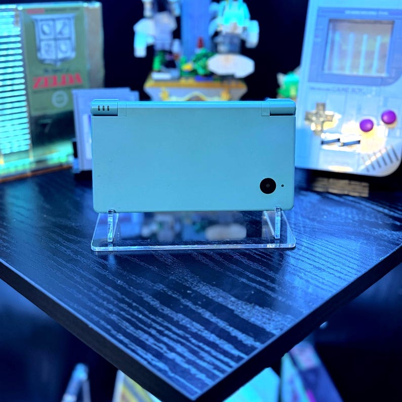 May include: A light teal Nintendo DS handheld game console on a clear acrylic stand. The console is closed, showing the top screen and buttons. Other retro gaming items are visible in the background, including a Game Boy.