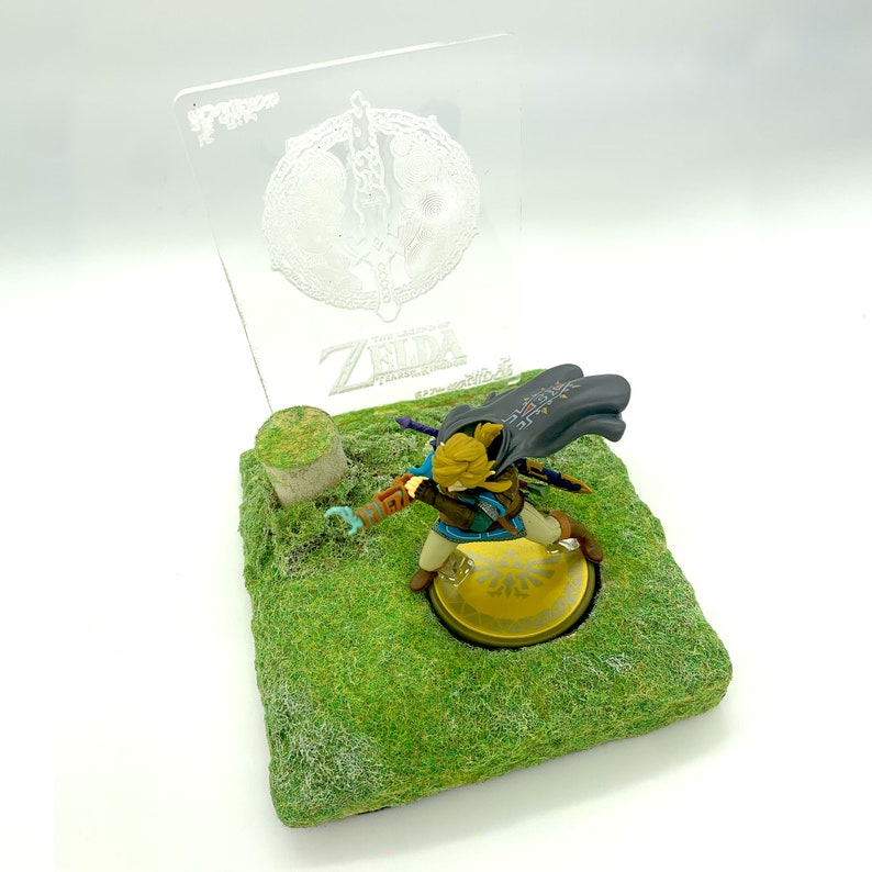 May include: A figurine of a character from The Legend of Zelda: Tears of the Kingdom, posed on a grassy base with a clear acrylic backdrop. The backdrop features the game's logo and a circular emblem. The figurine is in action, with a sword and shield.