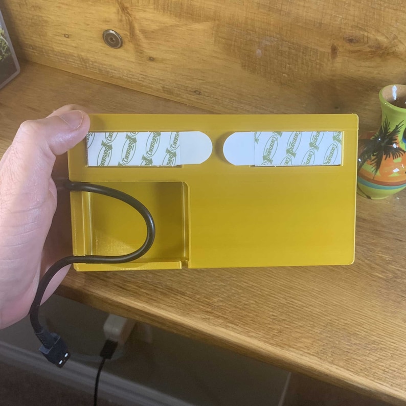 May include: A yellow plastic wall mount with two adhesive strips, a small compartment, and a cable management slot. The mount is designed to hold a small electronic device.