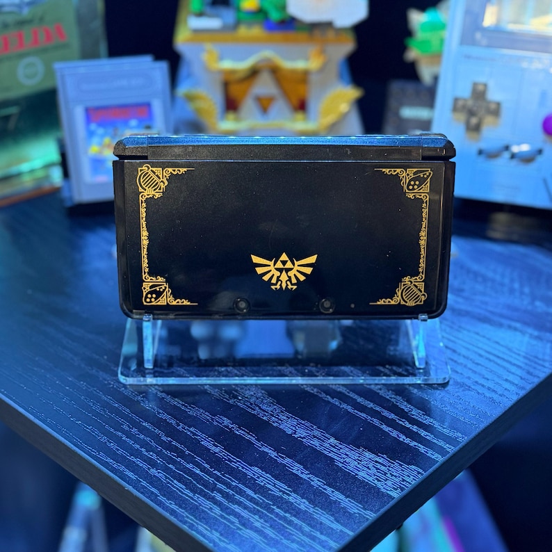 May include: Black handheld gaming console with gold decorative accents and a gold emblem. The console is displayed on a clear acrylic stand. Other gaming accessories are visible in the background.