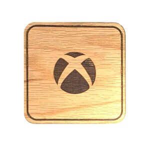 Op de afbeelding: Een houten onderzetter met een donkerbruin Xbox-logo in het midden. De onderzetter is vierkant en heeft een lichtbruine houtnerf.
