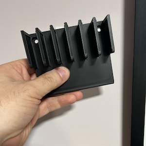 May include: A black, 3D-printed wall-mounted organizer with multiple vertical slots. The organizer is held by a hand and features a rectangular base with several evenly spaced dividers, and two holes for mounting. The object is made of plastic.