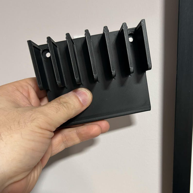 May include: A black, 3D-printed wall-mounted organizer with multiple vertical slots. The organizer is held by a hand and features a rectangular base with several evenly spaced dividers, and two holes for mounting. The object is made of plastic.