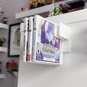 Puede incluir: Un soporte blanco montado en la pared que muestra cuatro cajas de juegos de Nintendo 3DS. Los juegos incluyen "Pok&eacute;mon Y", "Pok&eacute;mon Ultra Moon" y "Pok&eacute;mon Moon". La caja de "Pok&eacute;mon Moon" presenta un dise&ntilde;o p&uacute;rpura y blanco con el logotipo del juego.