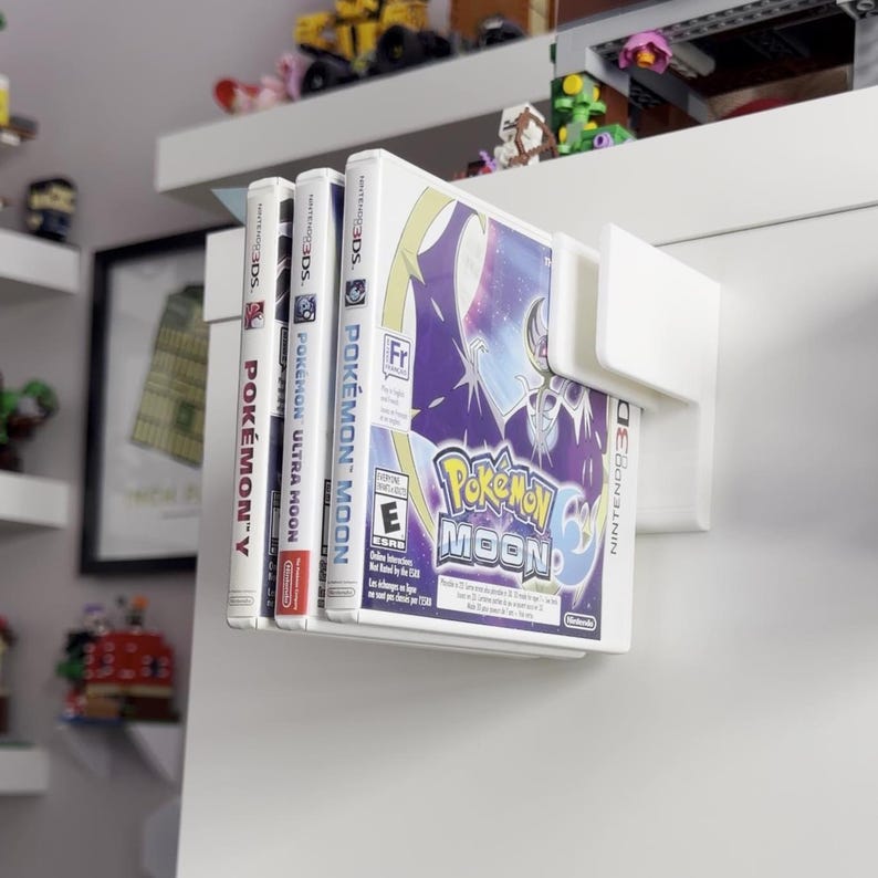 Puede incluir: Un soporte blanco montado en la pared que muestra cuatro cajas de juegos de Nintendo 3DS. Los juegos incluyen "Pok&eacute;mon Y", "Pok&eacute;mon Ultra Moon" y "Pok&eacute;mon Moon". La caja de "Pok&eacute;mon Moon" presenta un dise&ntilde;o p&uacute;rpura y blanco con el logotipo del juego.