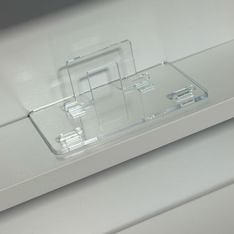 May include: A clear acrylic phone stand with a rectangular base and a vertical support. The stand has a minimalist design and is suitable for holding a phone or tablet. The stand is designed to be placed on a flat surface.