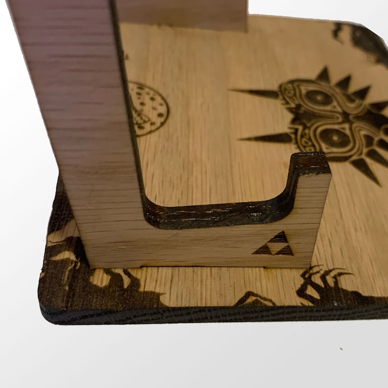 May include: Wooden stand with a cutout for a video game cartridge. The stand is decorated with a wood-burned image of Majora's Mask from the Legend of Zelda video game series.
