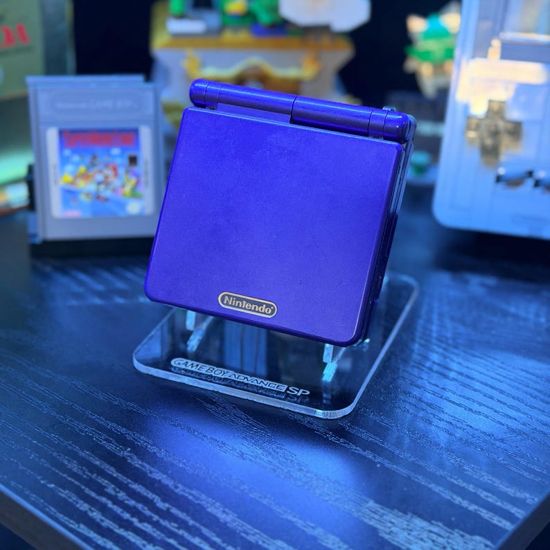 May include: A purple Nintendo Game Boy Advance SP handheld console on a clear acrylic stand. The stand is labeled "Game Boy Advance SP". A Game Boy cartridge is visible in the background.
