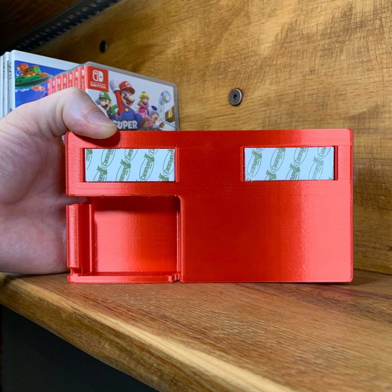 May include: Red plastic storage case with two compartments for video game cartridges. The case has Command brand adhesive strips on the back for mounting.
