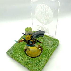 May include: A figurine of Link from The Legend of Zelda: Tears of the Kingdom, posed on a grassy base with a clear acrylic backdrop. The base has a gold emblem. The backdrop features the game's logo and a design.