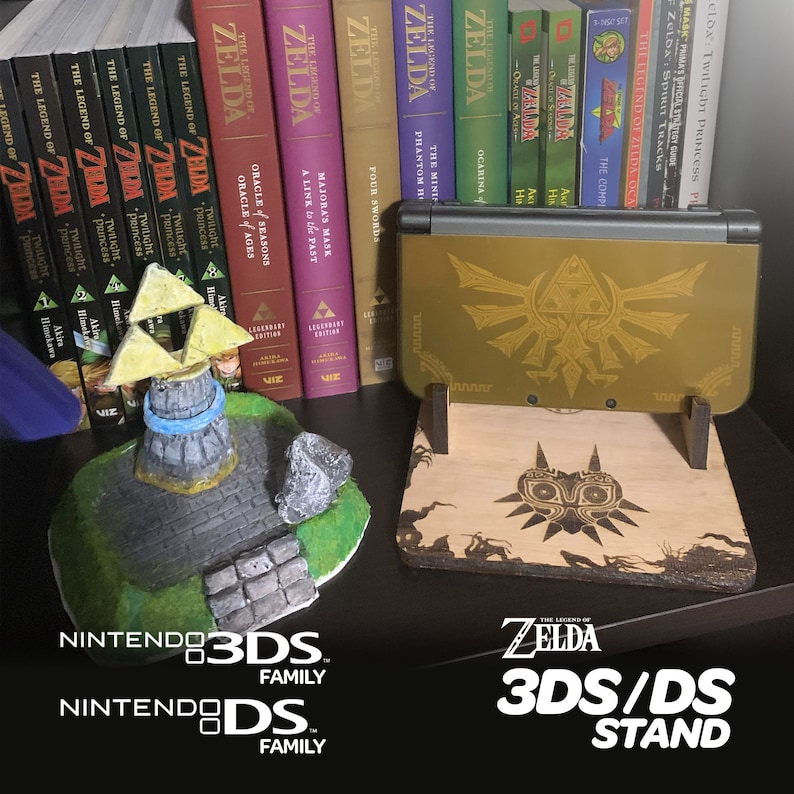 May include: A wooden stand for a Nintendo 3DS or DS handheld gaming console. The stand is decorated with a wood-burned image of the Triforce and Majora's Mask from the Legend of Zelda video game series. The stand is sitting on a shelf with a collection of Legend of Zelda video game cartridges.