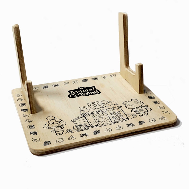 May include: A wooden stand with two upright supports, designed to hold video game cartridges. The stand features a printed design of characters from the Animal Crossing video game series, including the text "Animal Crossing" and " Nook's Cranny".