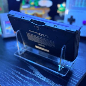 May include: A black Nintendo DS XL handheld game console is displayed on a clear acrylic stand. The console has a blue top and the Nintendo DS logo is visible. The stand is on a dark surface.