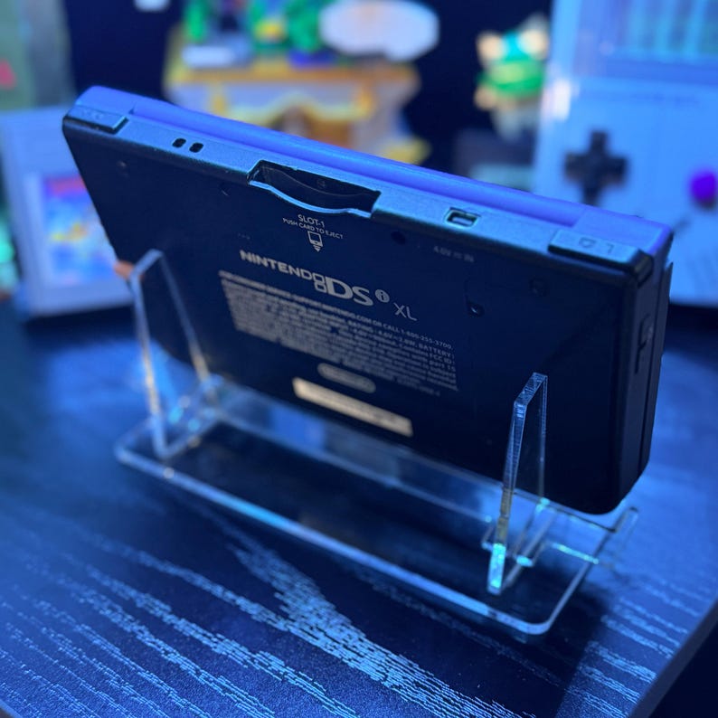 May include: A black Nintendo DS XL handheld game console is displayed on a clear acrylic stand. The console has a blue top and the Nintendo DS logo is visible. The stand is on a dark surface.