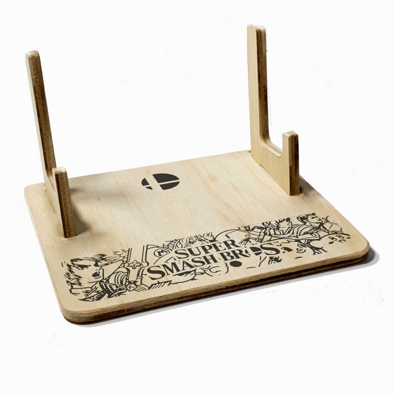 May include: A wooden stand with two upright supports for displaying video game cartridges. The stand is decorated with a black and white illustration of characters from the Super Smash Bros. video game series.