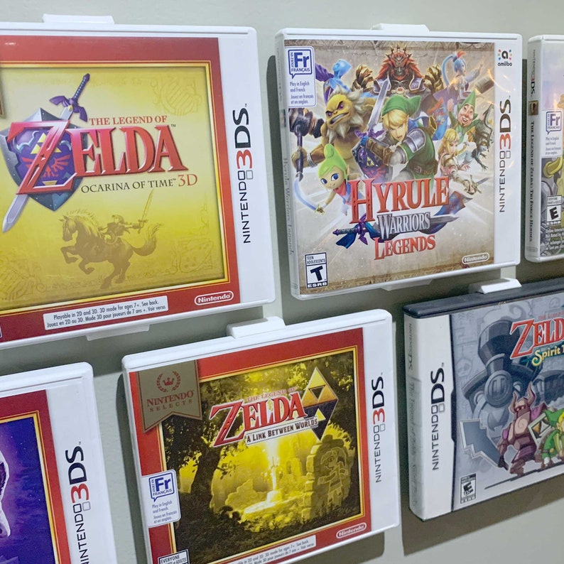 May include: A collection of six Nintendo 3DS game cartridges, including The Legend of Zelda: Ocarina of Time 3D, Hyrule Warriors Legends, The Legend of Zelda: A Link Between Worlds, and The Legend of Zelda: Spirit Tracks. The cartridges are displayed on a white wall.
