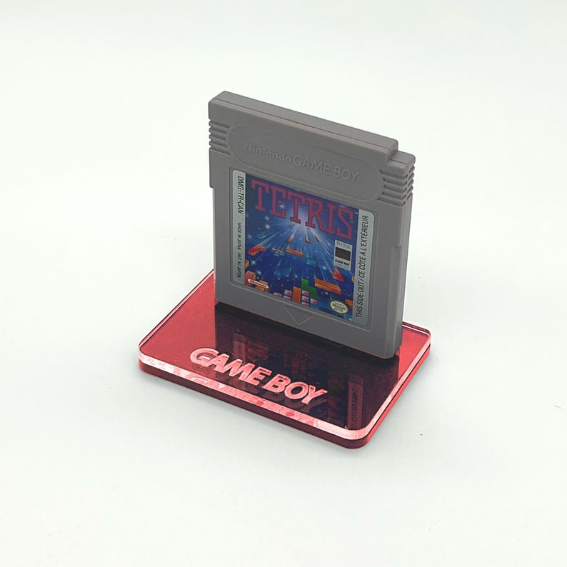 May include: A red acrylic Game Boy stand with the words "GAME BOY" printed on the front. A gray Game Boy cartridge with the game "TETRIS" is displayed on the stand.