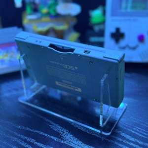 May include: A light blue Nintendo DS handheld game console is displayed on a clear acrylic stand. The console is angled, showcasing the top edge with the Nintendo DS logo. The stand is rectangular with a base and two vertical supports.