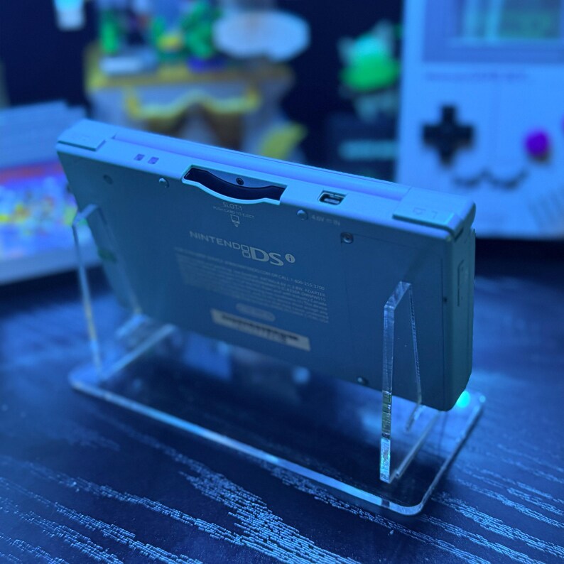 May include: A light blue Nintendo DS handheld game console is displayed on a clear acrylic stand. The console is angled, showcasing the top edge with the Nintendo DS logo. The stand is rectangular with a base and two vertical supports.