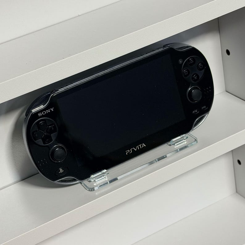 May include: A black Sony PlayStation Vita handheld game console, resting on a clear plastic stand. The console has a black screen and buttons, with the word "SONY" above the directional pad and "PS VITA" below the screen.