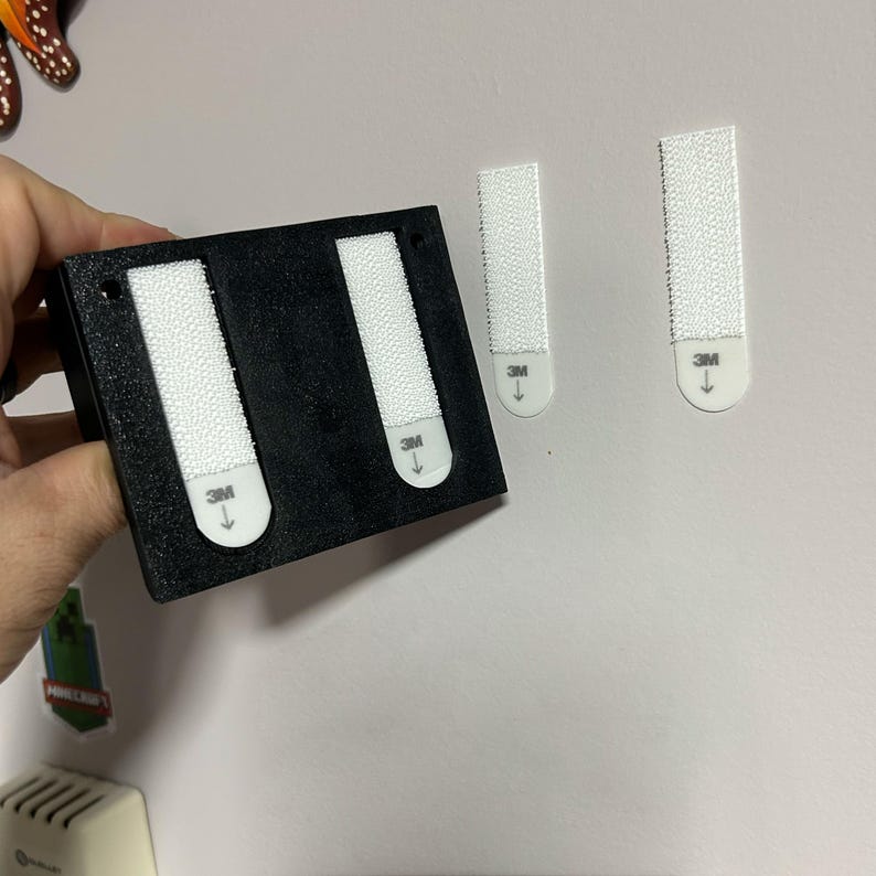 May include: A black rectangular object with two white adhesive strips attached, along with two separate white adhesive strips. The strips have the "3M" logo and an arrow. The object is likely for mounting or hanging items.