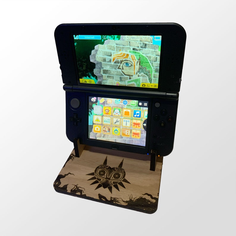 May include: A black Nintendo 3DS handheld game console is displayed on a wooden stand. The top screen shows a video game scene with a green and brown color scheme. The bottom screen displays a menu of game icons. The stand features a carved design.