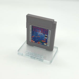 May include: A gray Game Boy cartridge for the game Tetris is displayed on a clear acrylic stand. The stand has the word "GAMEBOY" engraved on it.