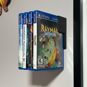 May include: Four video game cartridges for the Playstation Vita console. The cartridges are blue and black with colorful game titles and logos. The cartridges are displayed on a white wall with a black frame.