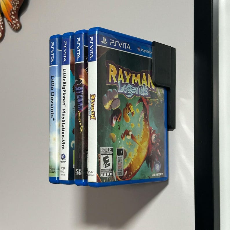 May include: Four video game cartridges for the Playstation Vita console. The cartridges are blue and black with colorful game titles and logos. The cartridges are displayed on a white wall with a black frame.